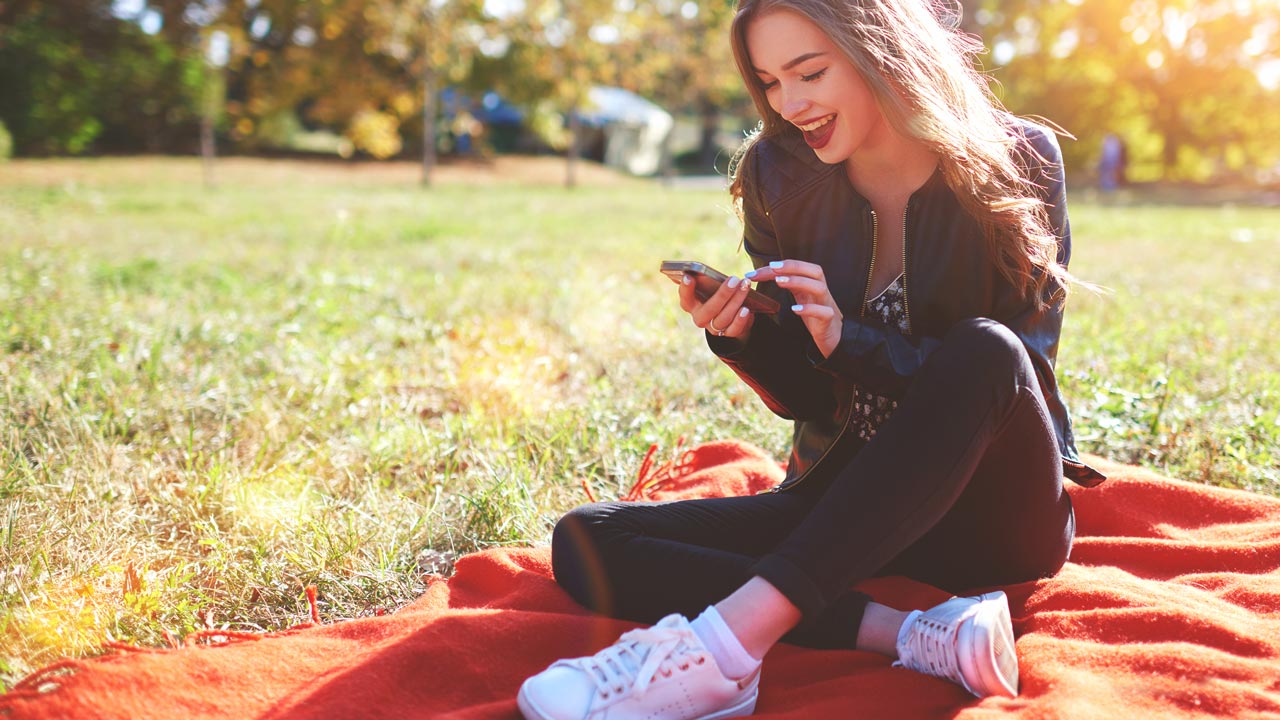 Junge Frau sitzt im Park auf einer Decke und schaut in ihr Handy