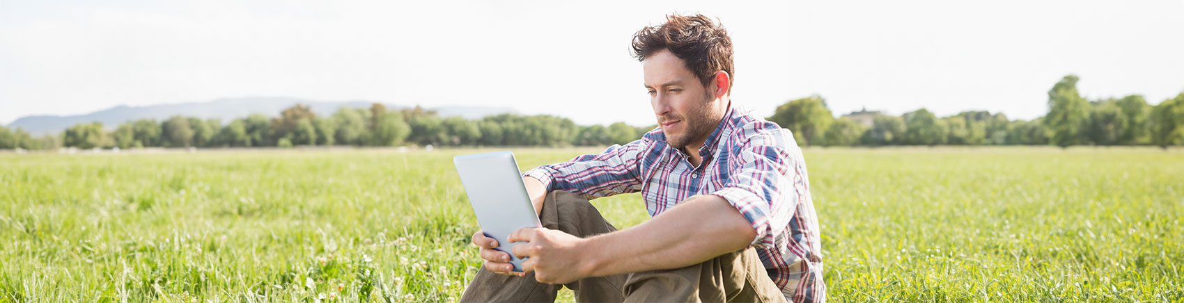 Junger Mann sitzt auf einer Wiese und schaut in sein Tablet