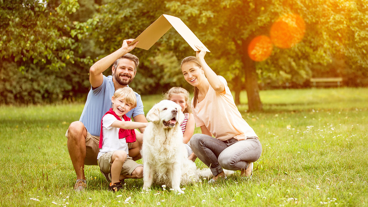 Vierköpfige Familie mit Hund hält ein Kartondach über Ihren Kopf als Symbol für ein Eigenheim